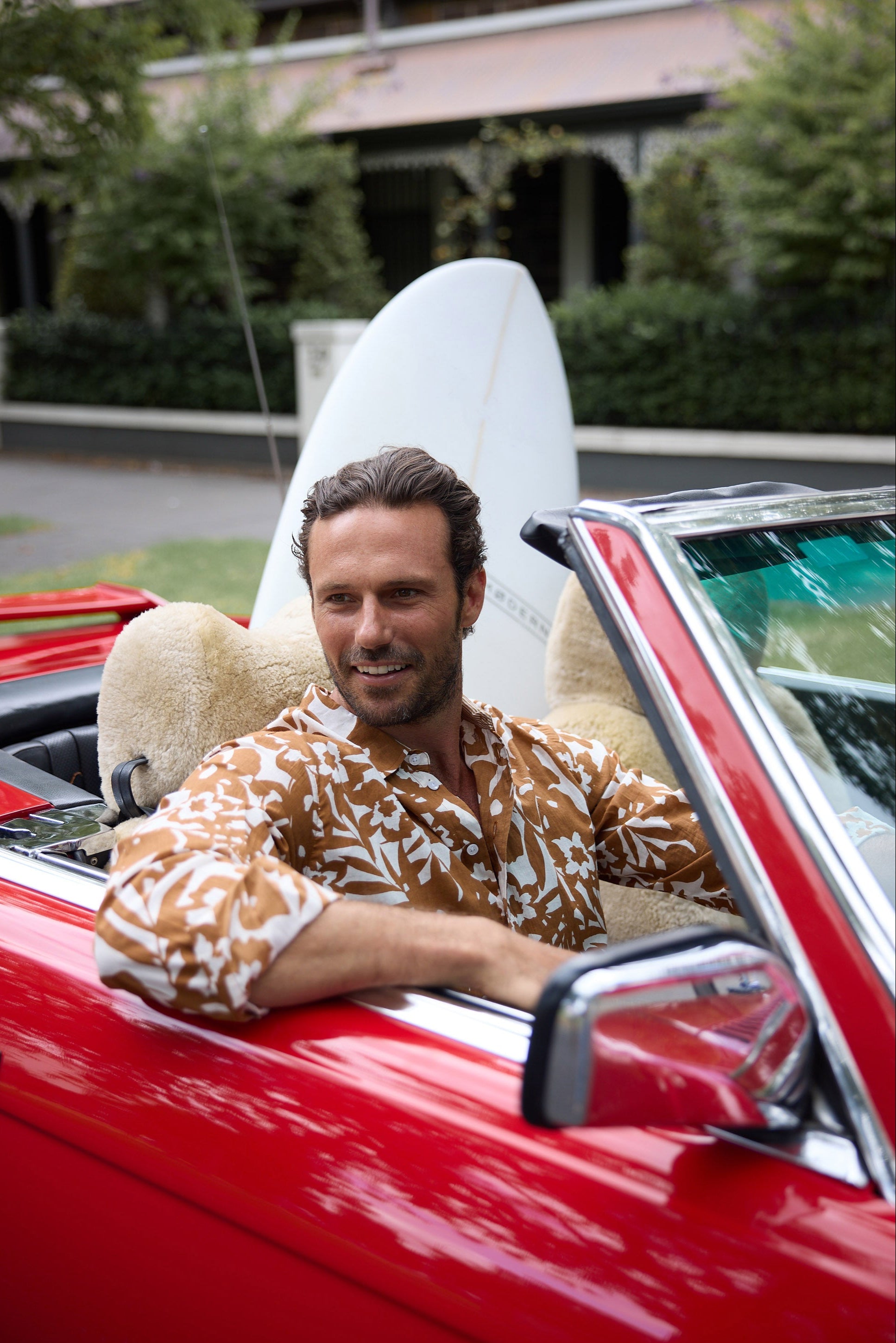 Man sitting in a red convertible with a surfboard on his lap, outdoors.