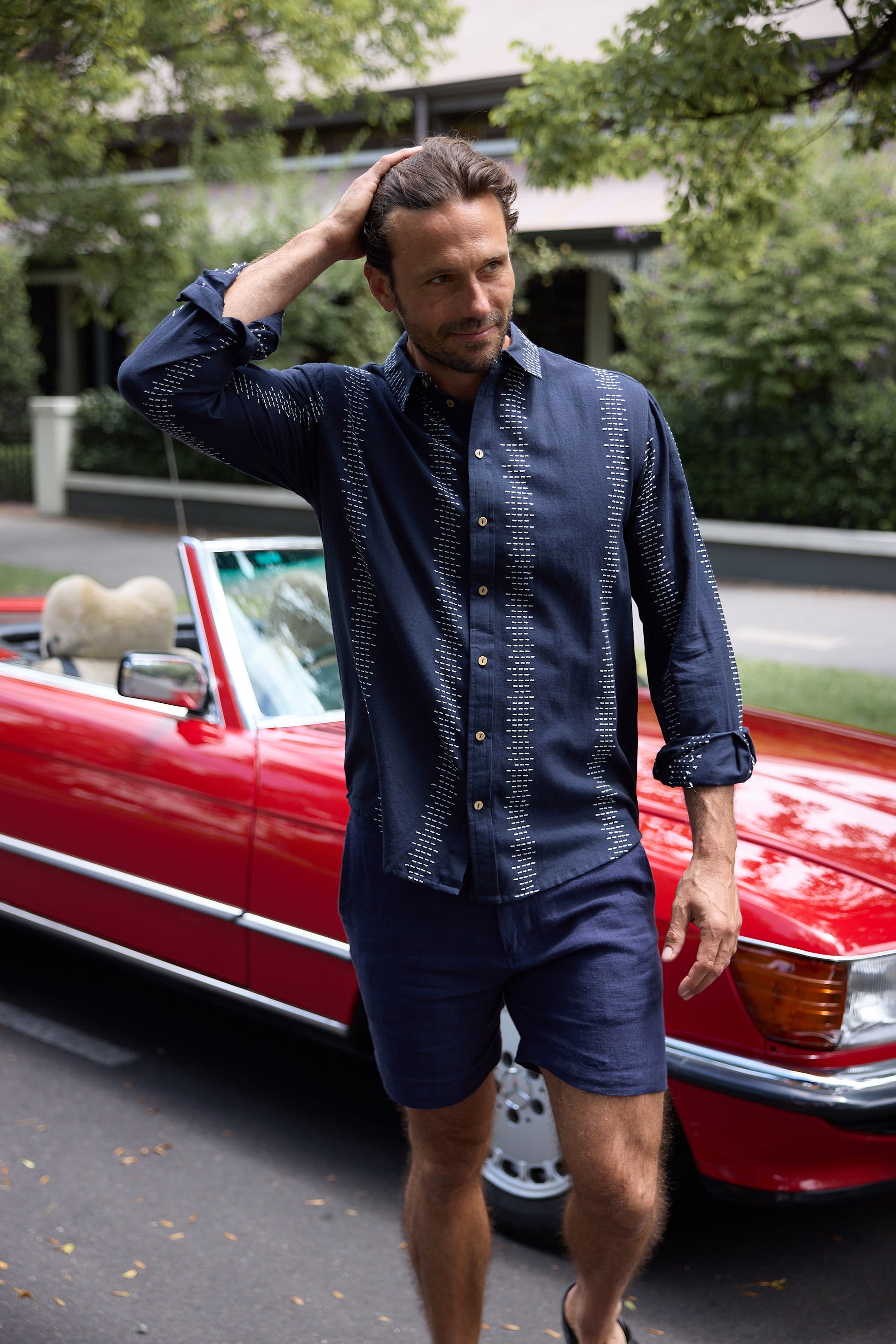 Man in navy shirt and shorts standing next to a red car with greenery in the background