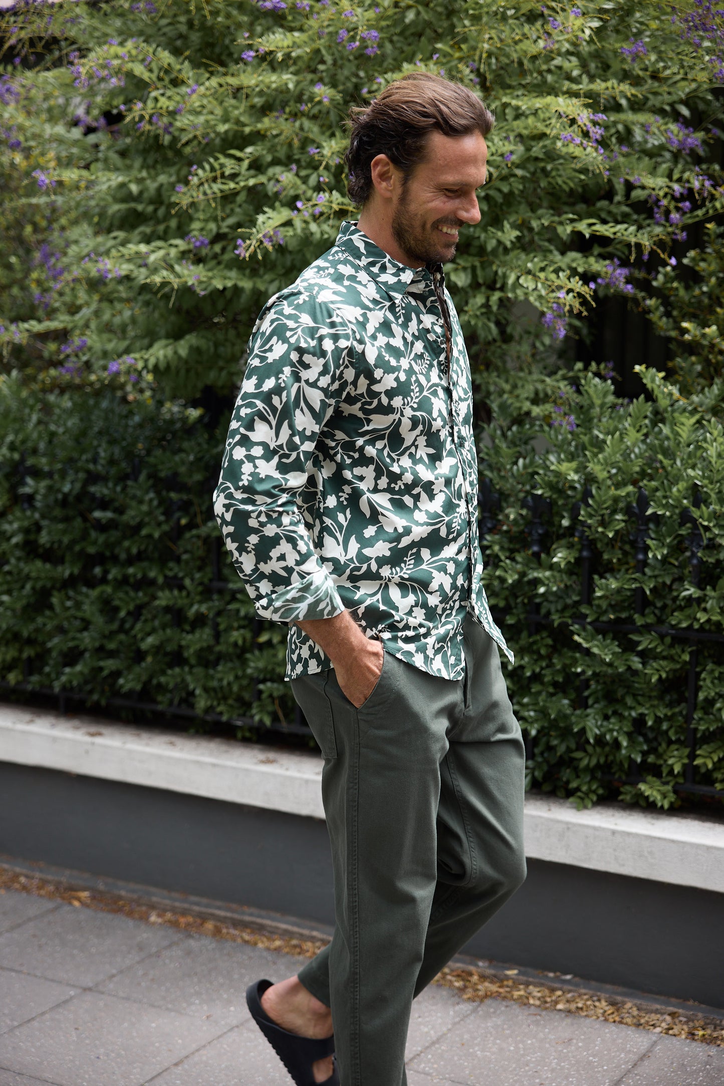 Man wearing a green patterned shirt and gray pants standing outdoors with greenery in the background