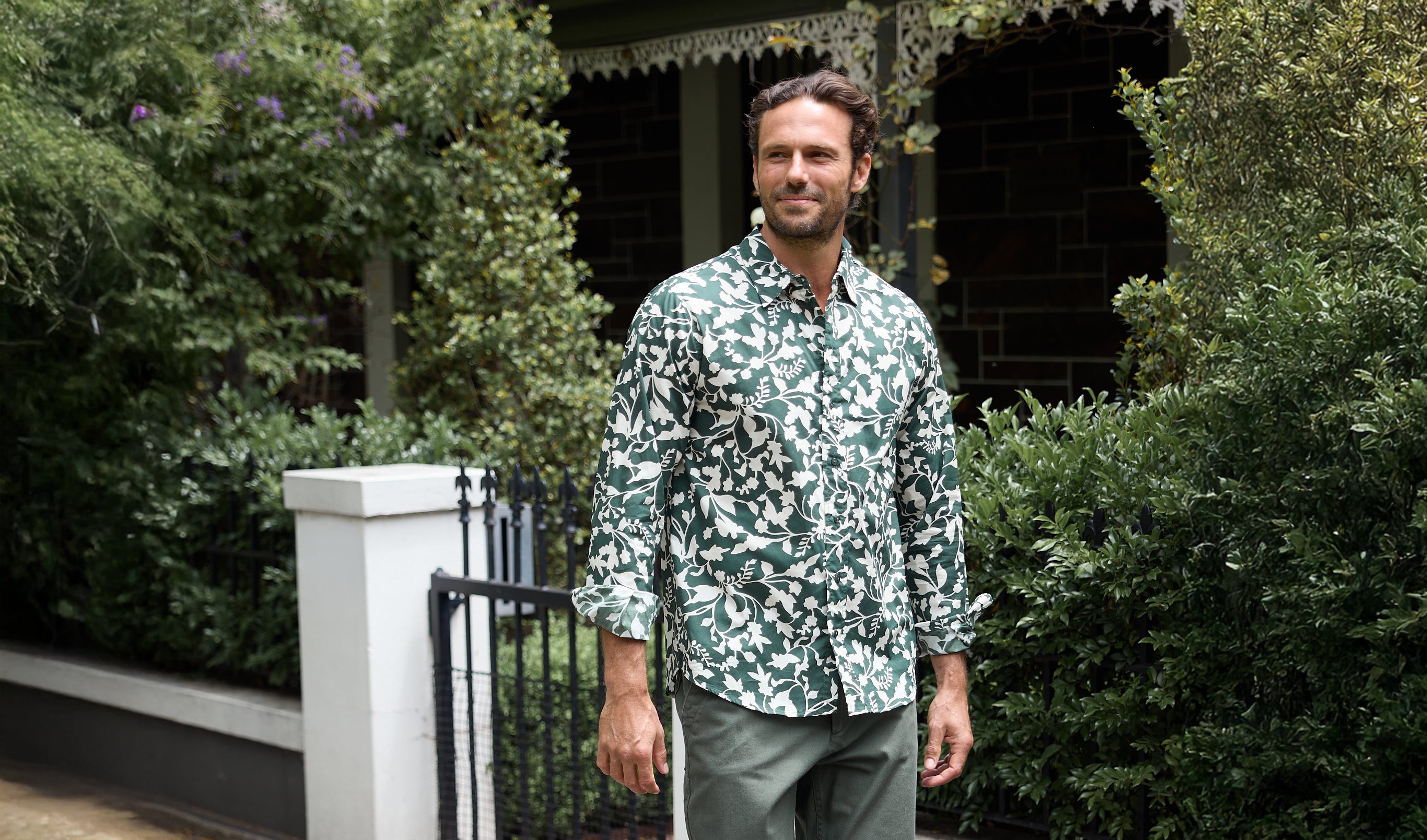 Man wearing a patterned shirt standing in front of a house with greenery.