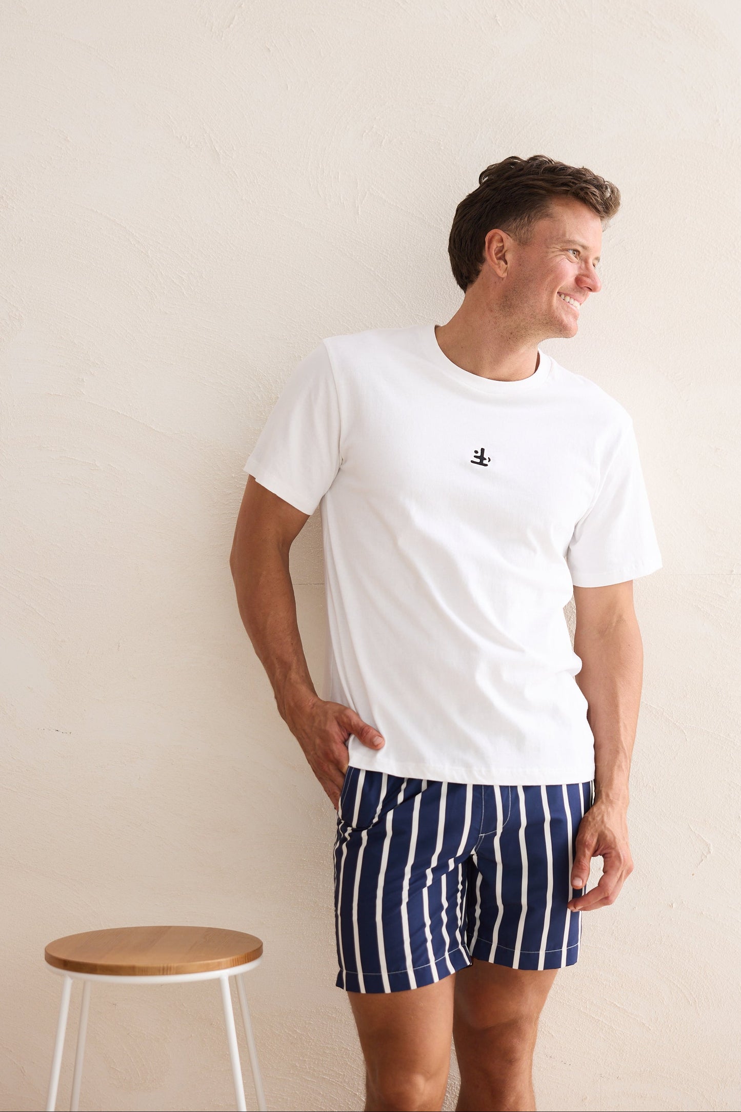 Man wearing a white t-shirt and navy striped shorts standing against a plain wall.