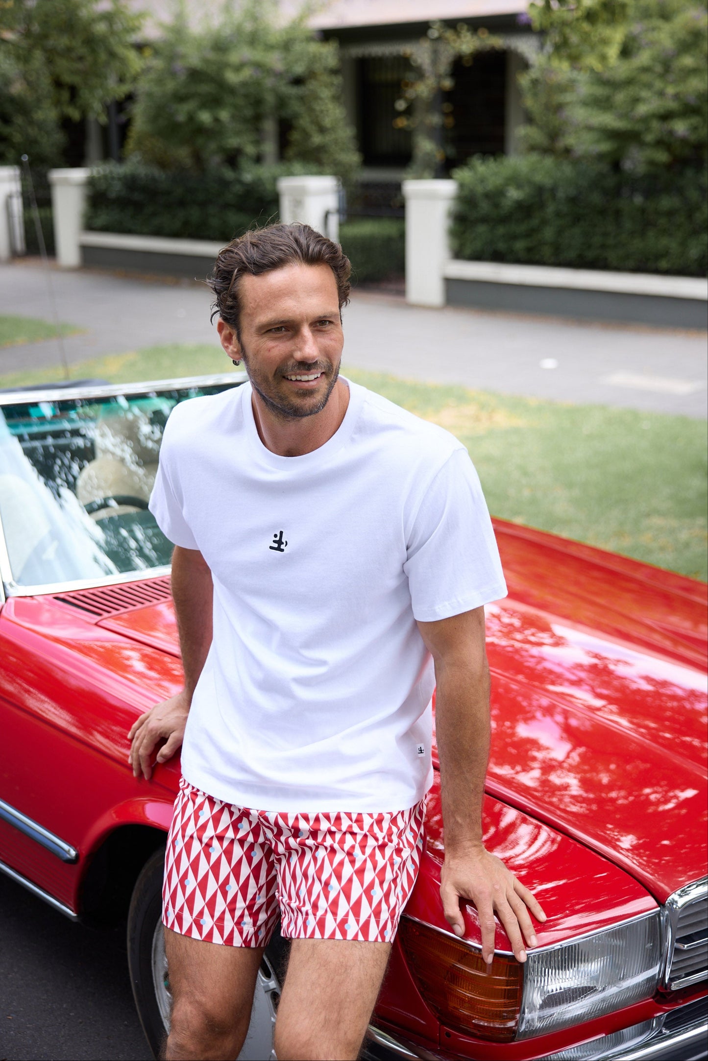 Man in a white t-shirt and red patterned shorts standing next to a red convertible car.