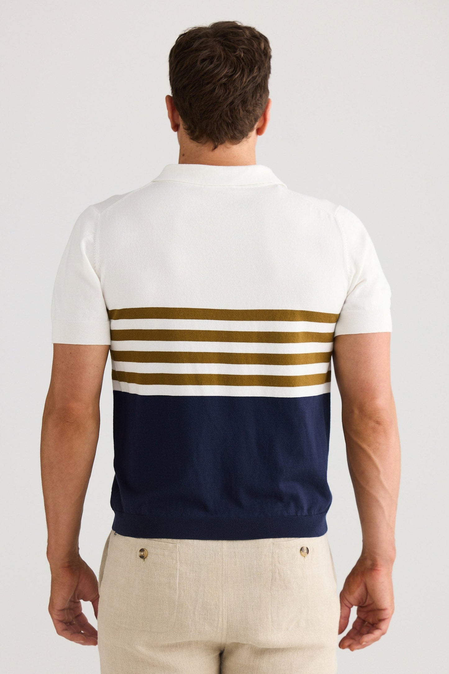 Man wearing a white polo shirt with blue and gold stripes and beige shorts on a light gray background