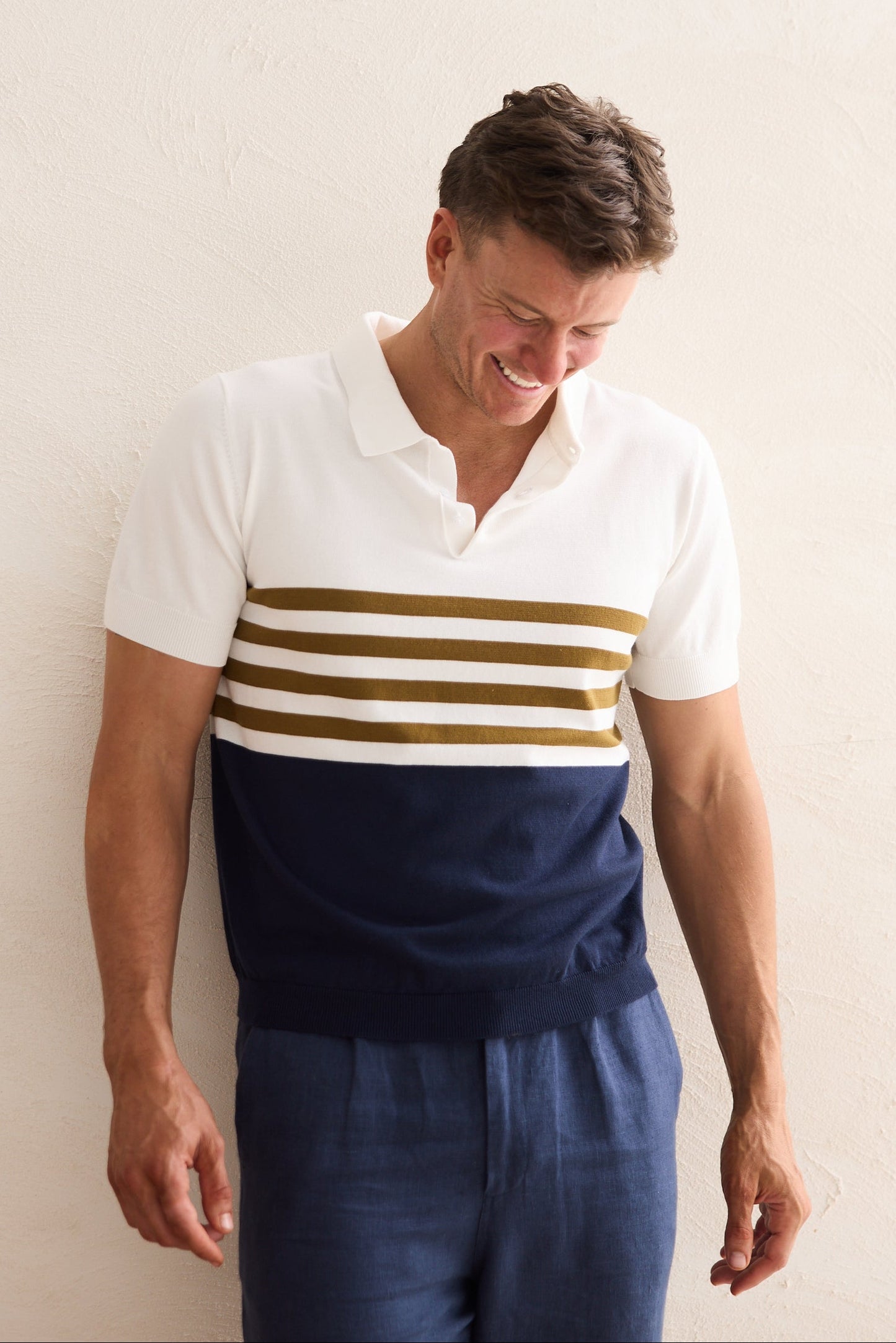 Man wearing a white polo shirt with navy and gold stripes and navy pants against a plain background