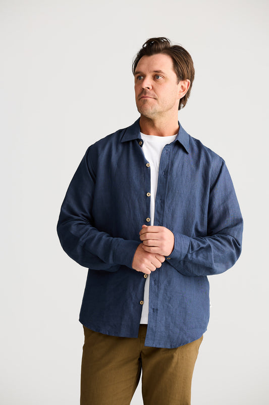 Man wearing a blue shirt over a white shirt with brown pants on a light gray background