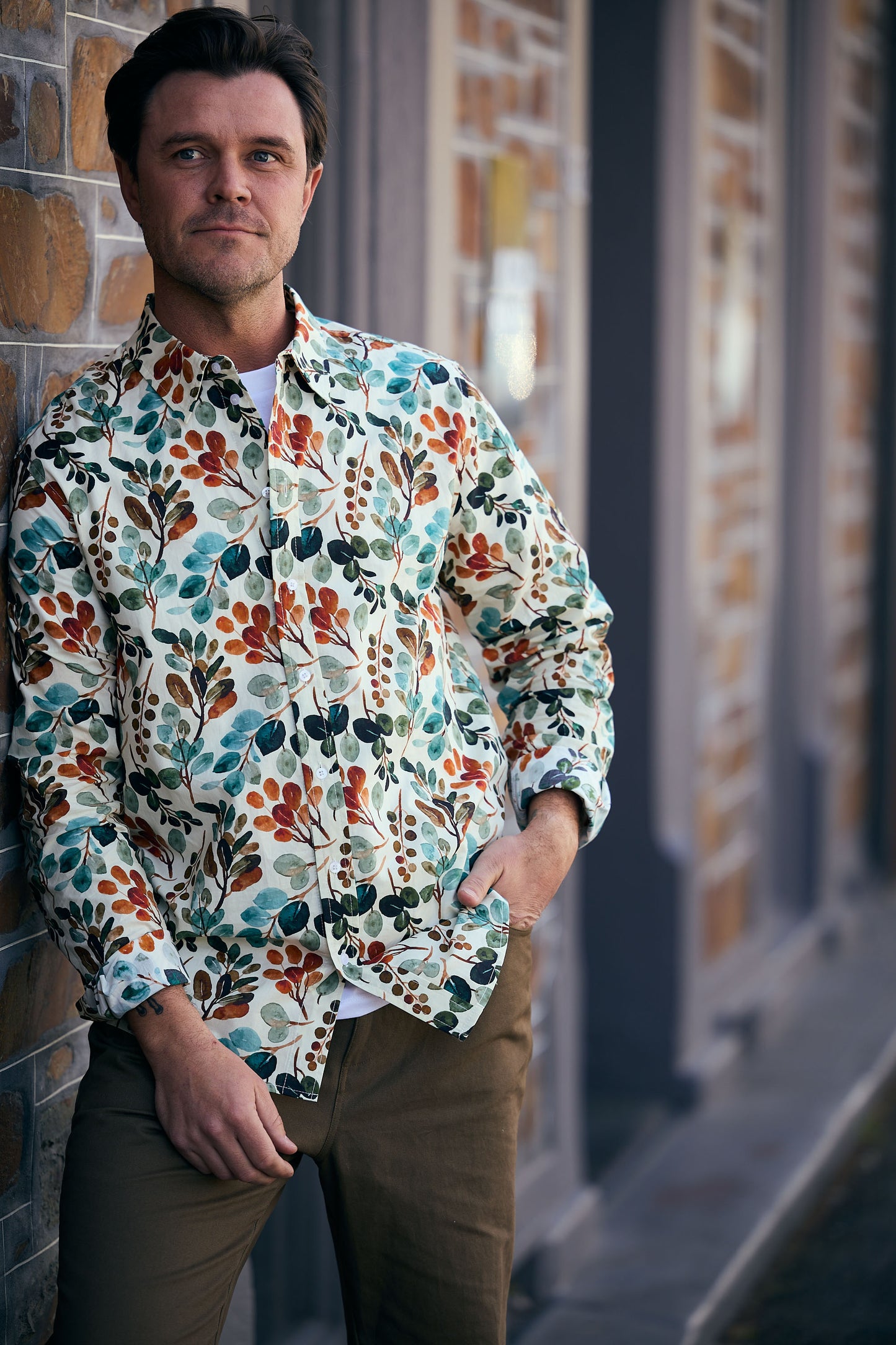 Man wearing a colorful floral shirt leaning against a brick wall.