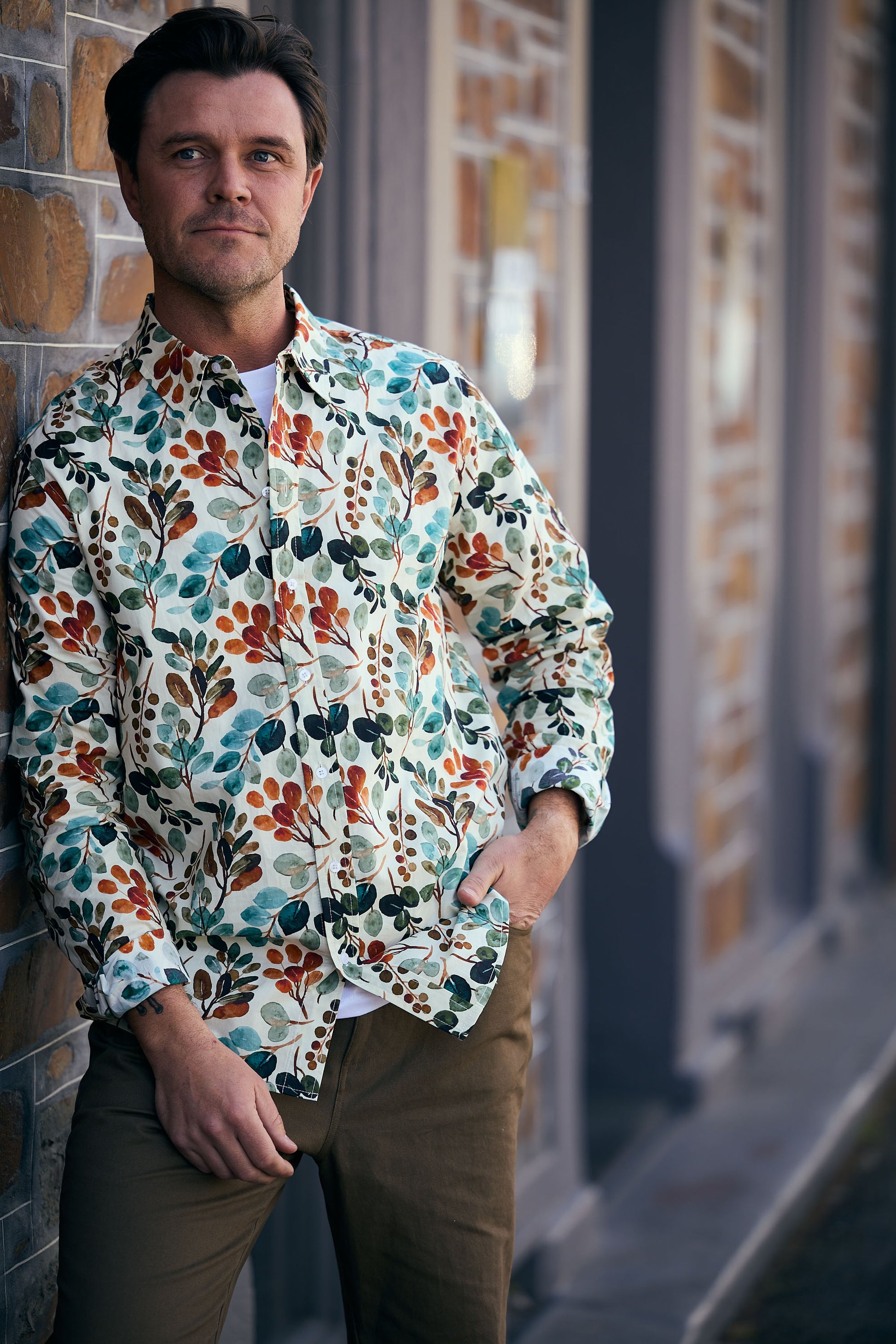 Man wearing a colorful floral shirt leaning against a brick wall.