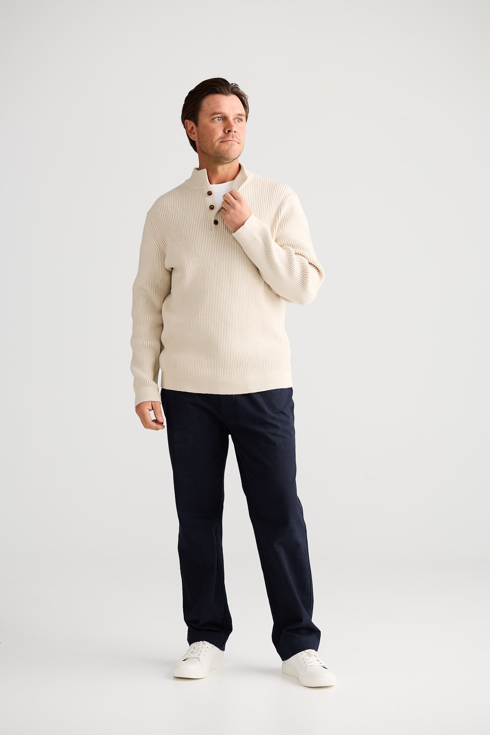 Man wearing a cream sweater and navy pants holding a coffee cup on a white background
