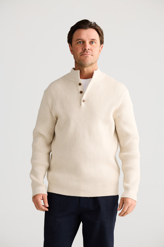 Man wearing a cream cardigan over a white shirt with navy pants on a light gray background