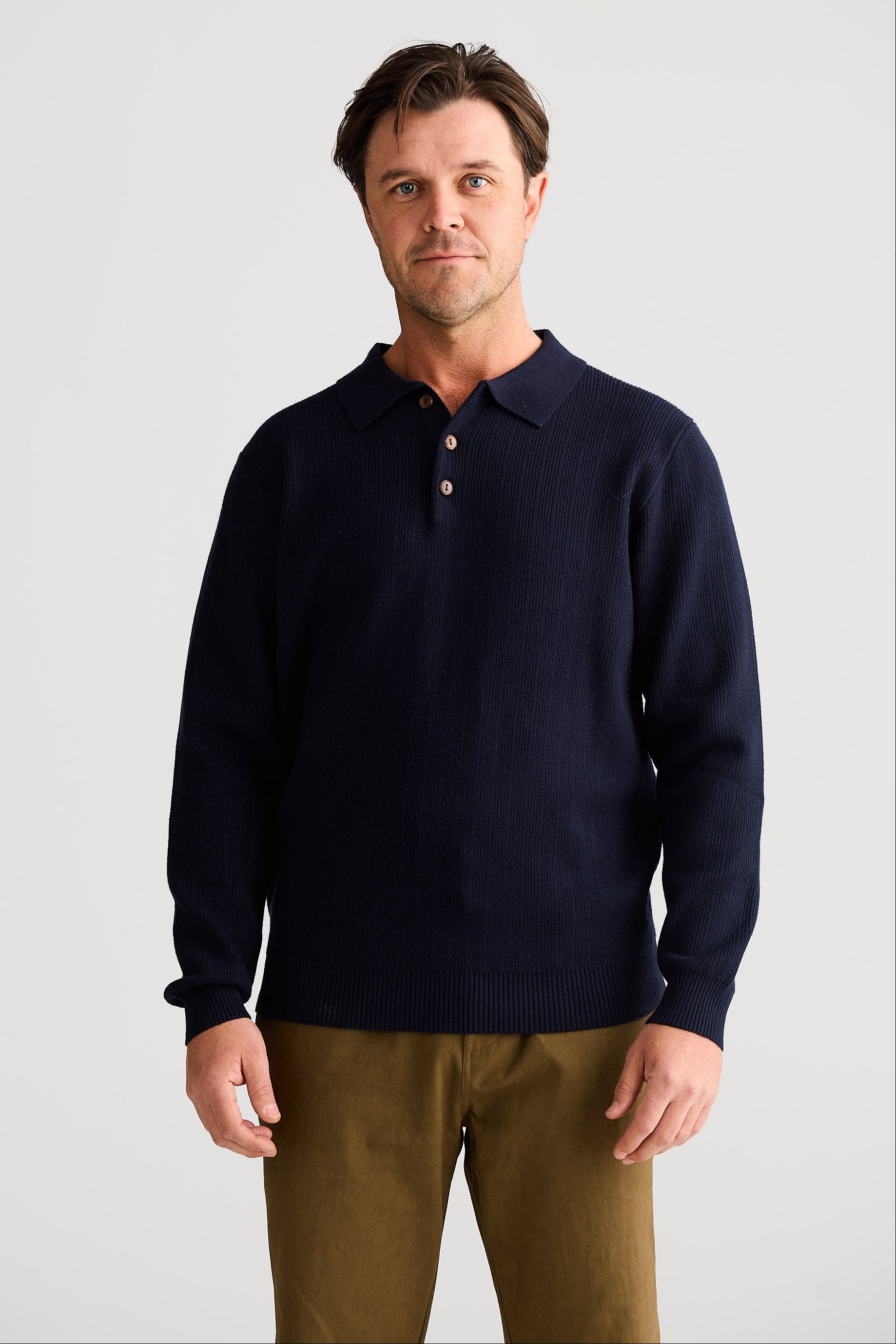 Man wearing a navy sweater over a collared shirt with brown pants on a light gray background